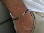 A close-up of a person's wrist wearing a blue cord bracelet with a silver metal bar engraved with Hebrew text.