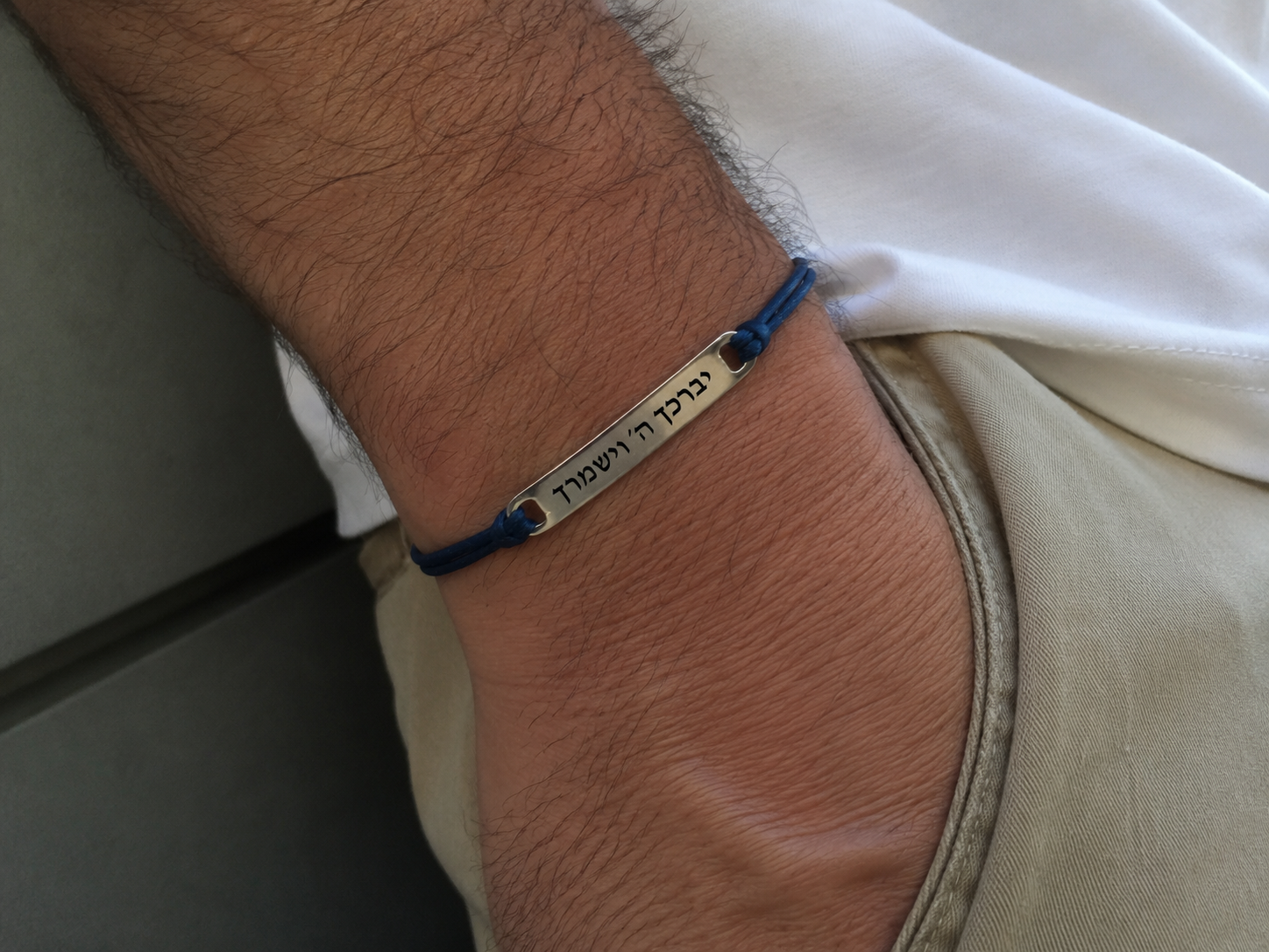 A close-up of a person's wrist wearing a blue cord bracelet with a silver metal bar engraved with Hebrew text.