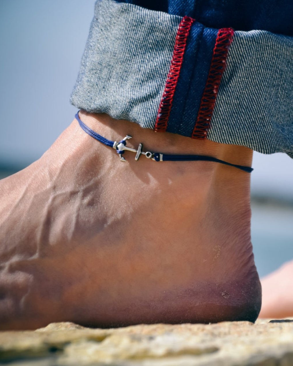 Anchor anklet for men on a foot with jeans rolled up, near water.