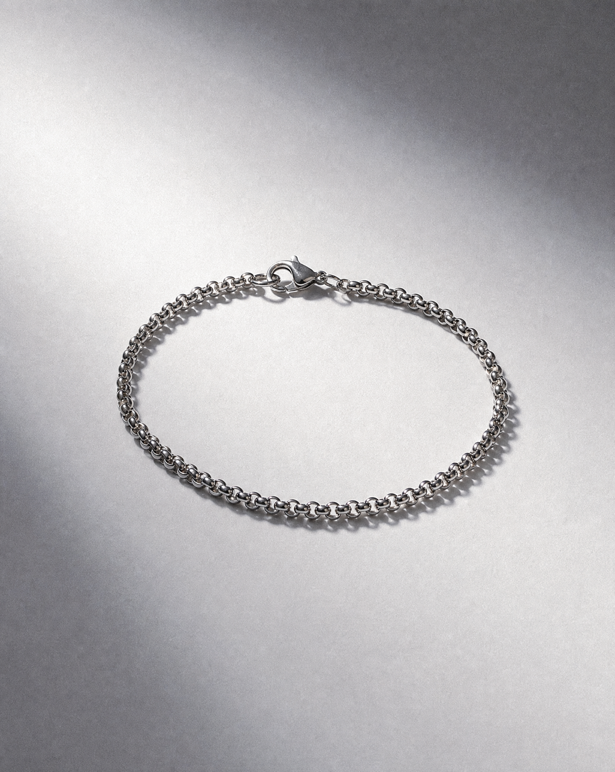 A silver-toned metal chain bracelet with a lobster clasp, displayed on a textured gray surface with dramatic lighting.