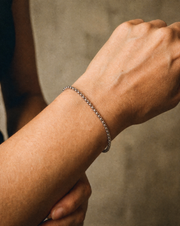 A close-up of a person's wrist wearing a delicate silver chain bracelet against a neutral textured background.