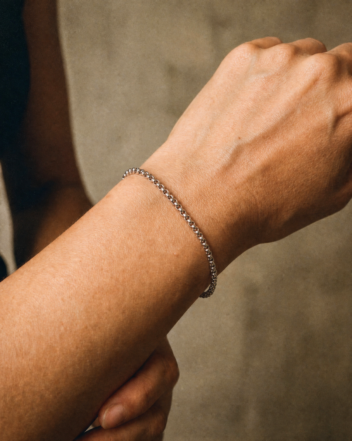 A close-up of a person's wrist wearing a delicate silver chain bracelet against a neutral textured background.