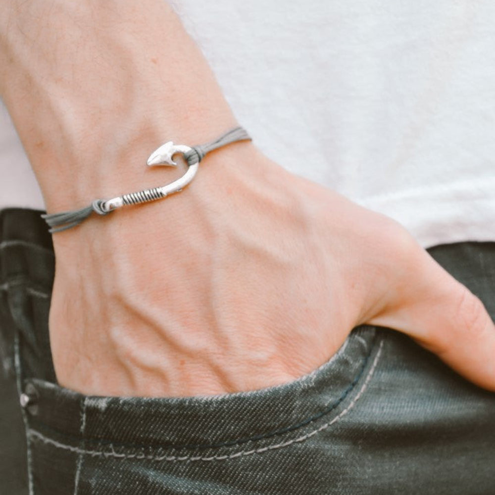 Hand wearing a silver bracelet with a fish hook charm on a plain background