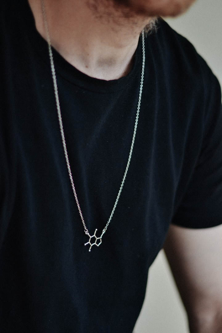 A man wearing a black t-shirt looking to the side, with a silver tone caffeine molecule pendant on a stainless steel chain necklace.