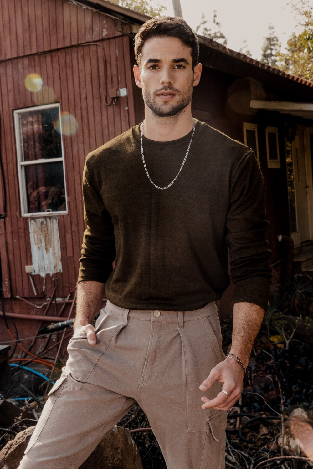 A man wearing a green sweater and brown pants, standing in front of a rustic backdrop.