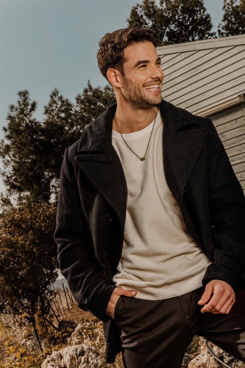 A man wearing a bronze Hamsa chain necklace, black jacket and a white shirt, standing outdoors with trees in the background.