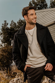 A man wearing a bronze Hamsa chain necklace, black jacket and a white shirt, standing outdoors with trees in the background.