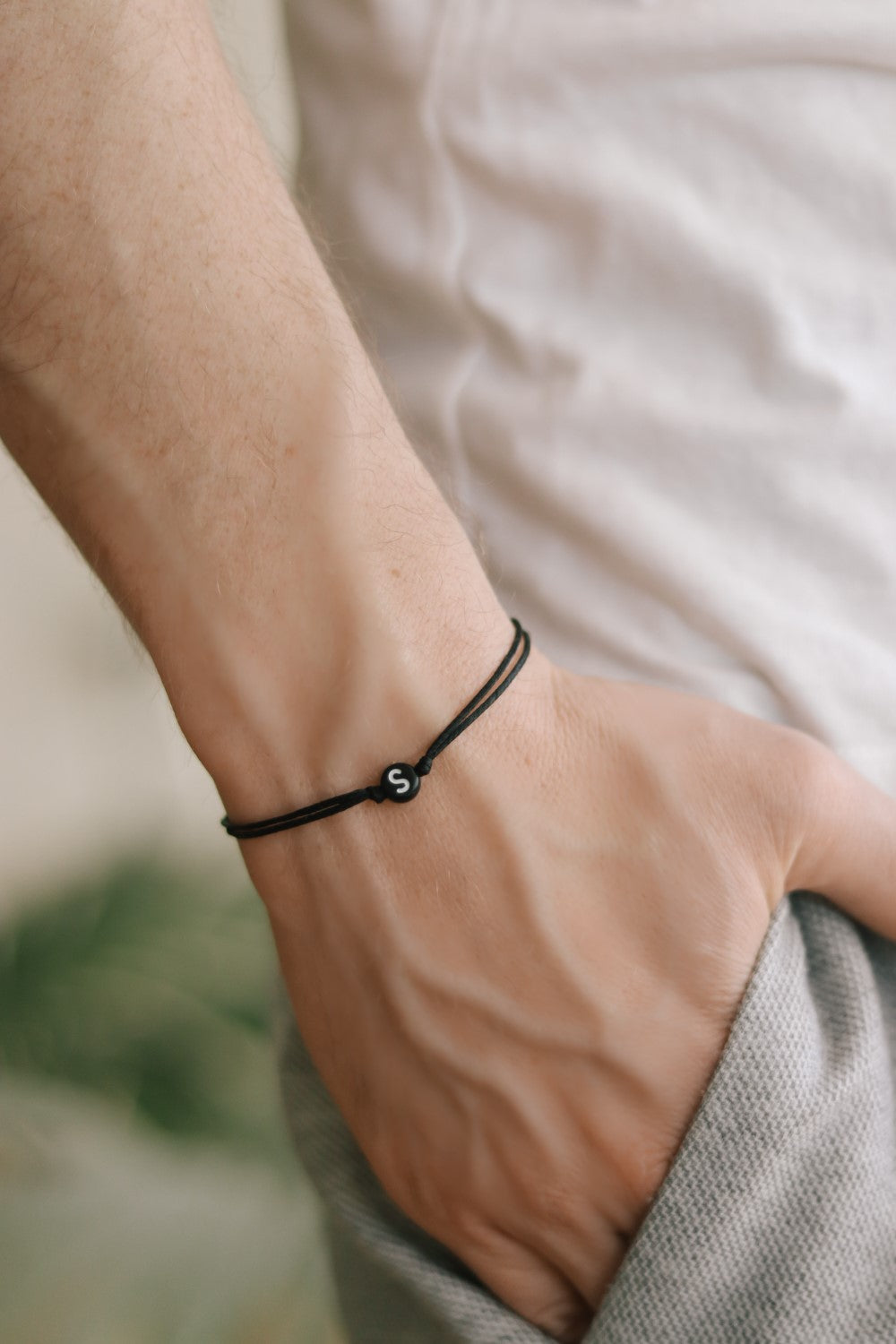 A man's wrist wearing a black cord bracelet with a black round bead and a initial letter