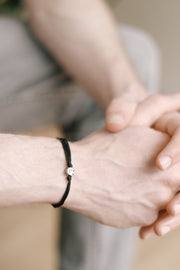 A close-up image of a man's wrist wearing a black cord bracelet with a silver plated bead charm