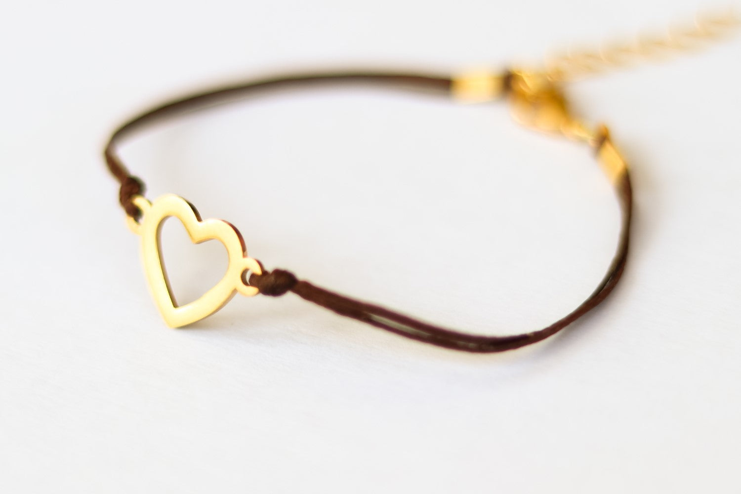 A gold heart charm bracelet with a dark brown cord and a gold-tone heart charm, displayed against a white background.